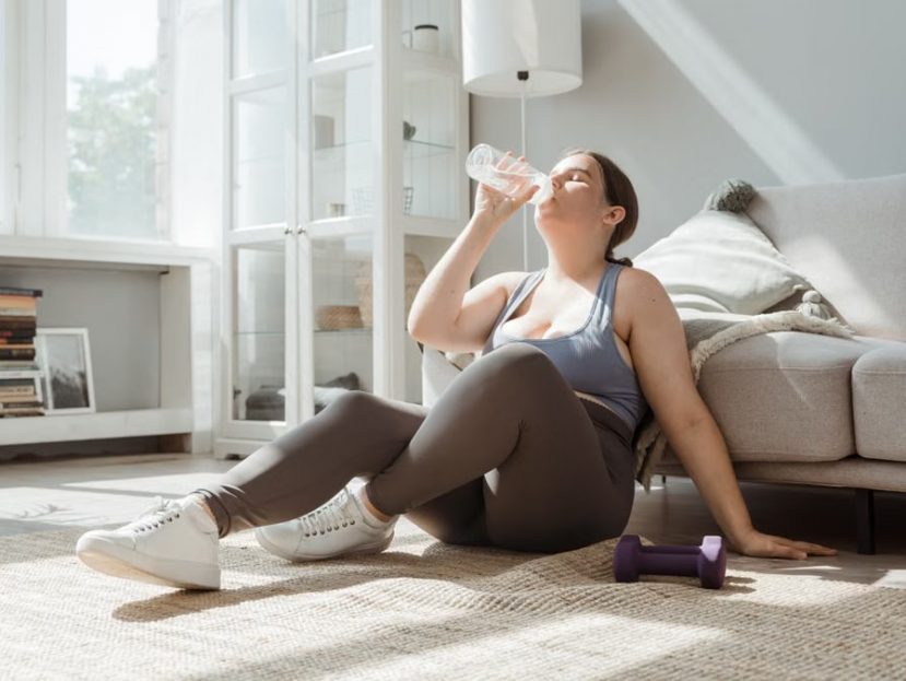 Woman sitting on living room floor, drinking water after workout, enjoying relaxation.